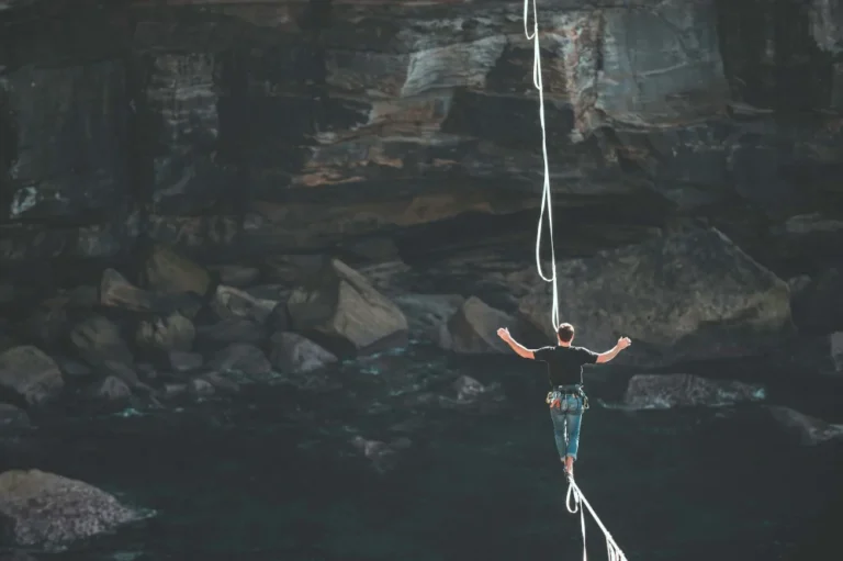 Bild eines Menschen, der über eine gespannte Highline einen Abgrund überwindet (mit Sicherung) als Sinnbild für den schmalen Grat von Perfektionismus