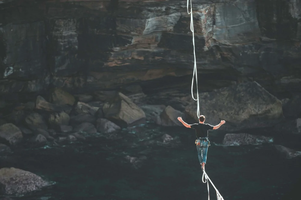 Bild eines Menschen, der über eine gespannte Highline einen Abgrund überwindet (mit Sicherung) als Sinnbild für den schmalen Grat von Perfektionismus